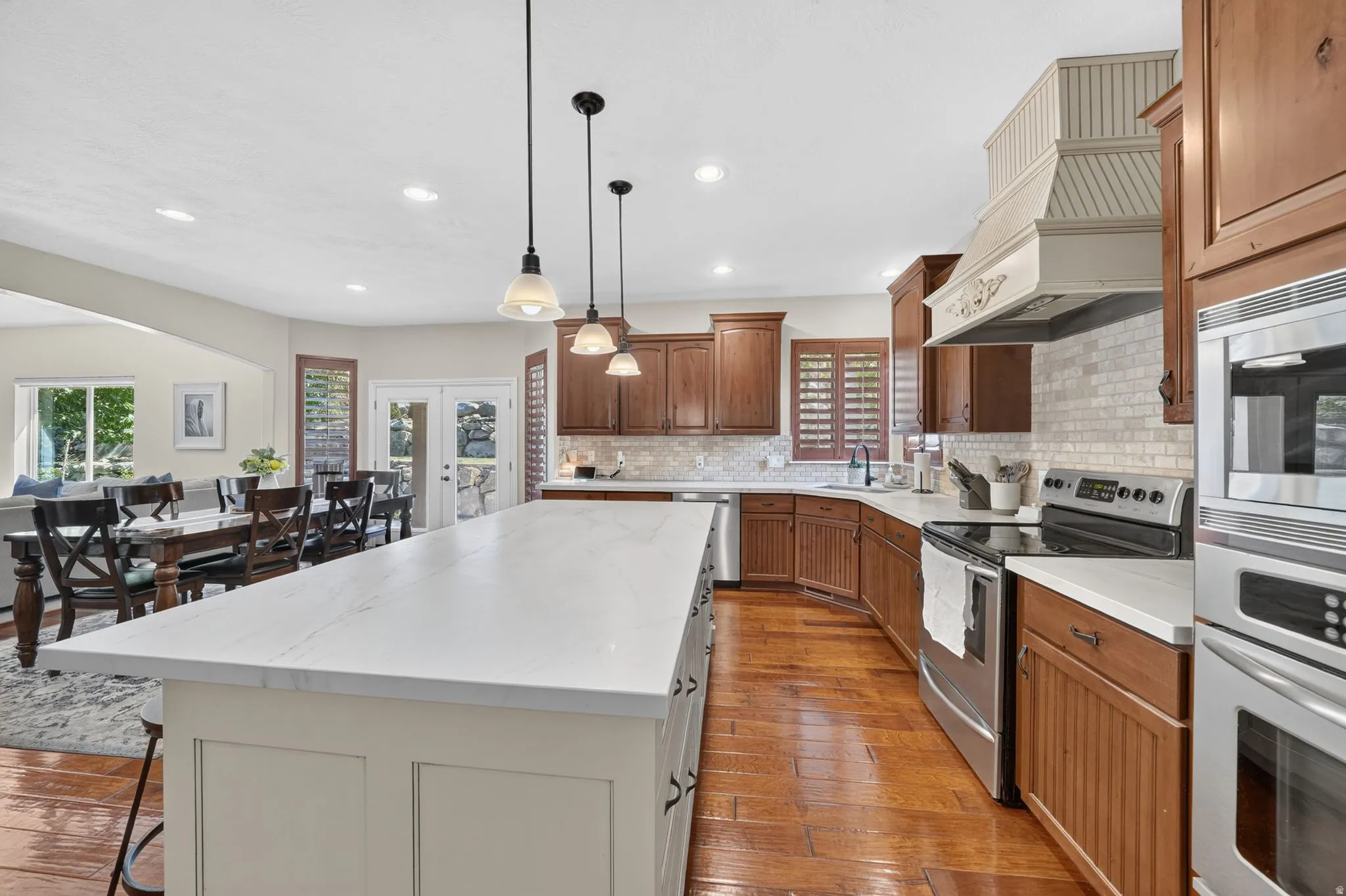 Expansive kitchen island with a white countertop, rich wood cabinetry, stainless steel appliances, decorative range hood, and tile backsplash