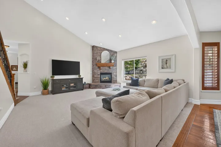 Vaulted ceiling with recessed lighting, stone-clad fireplace with wood mantel, hardwood flooring, and a large window with exterior greenery
