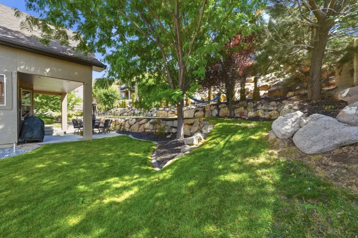 Expansive tiered backyard featuring a manicured lawn, concrete patio, covered outdoor living space, and stone retaining walls