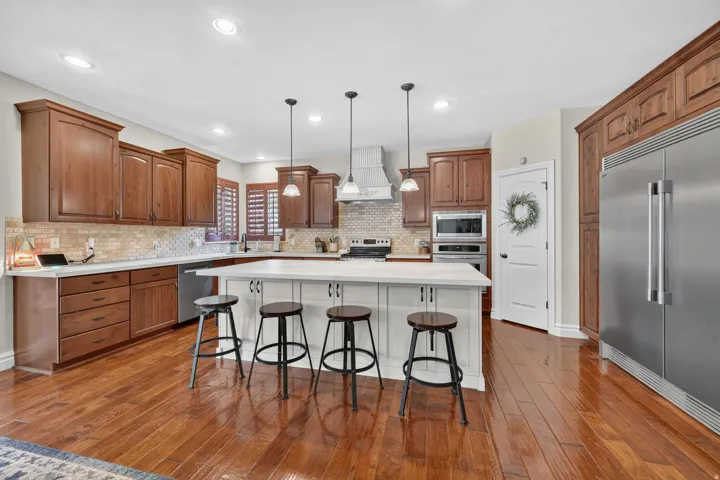 Wood-finish flooring, white kitchen island with pendant lighting, stainless steel appliances, wood cabinetry, and a tile backsplash