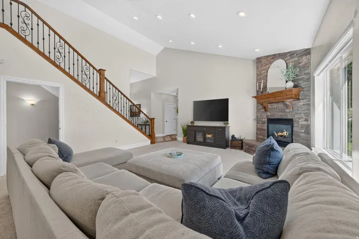 Living space featuring high ceilings, recessed lighting, a stone-clad fireplace with a wood mantel, a large window, and a staircase with wood handrails and wrought iron balusters