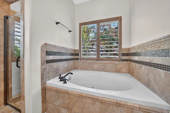 Built-in soaking tub with dark bronze fixtures, framed window with plantation shutters, tile surround with decorative accent band, and a glass-enclosed shower