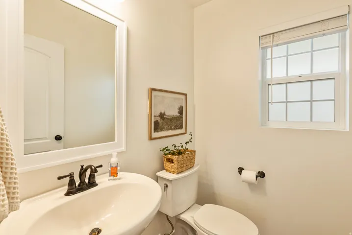 Bright bathroom featuring a curved pedestal sink with oil-rubbed bronze faucet, large framed mirror with integrated lighting, toilet with black hardware, and a window providing natural light
