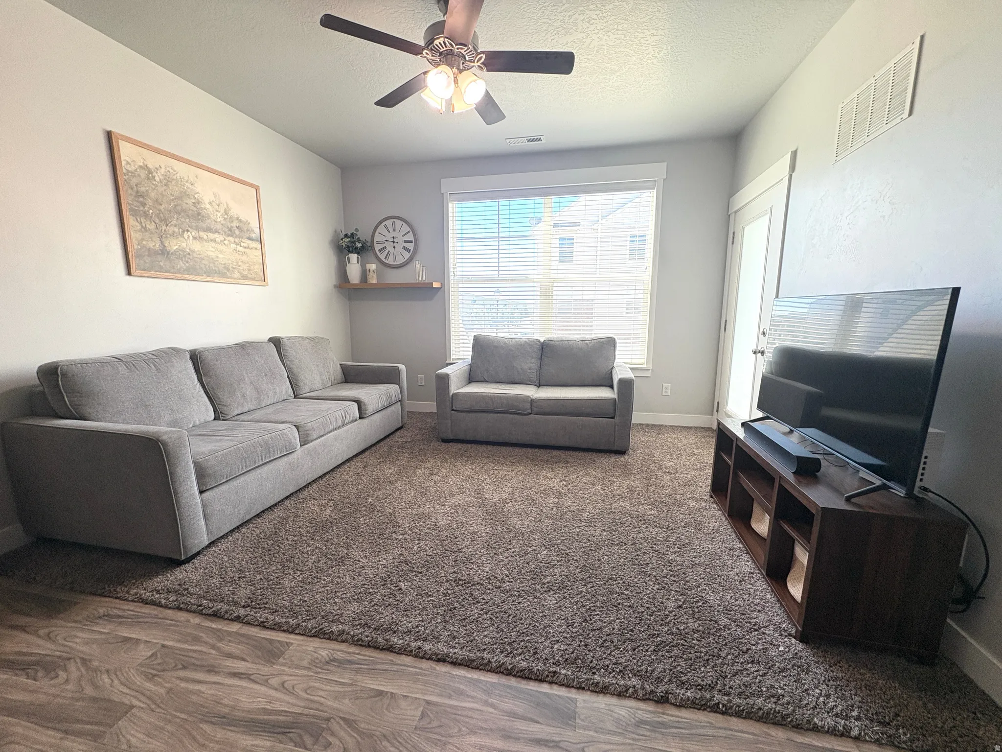 Living area featuring wood-finish flooring, a ceiling fan with integrated lighting, and a large window with blinds
