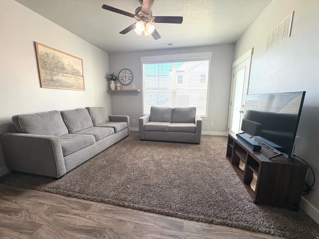 Living area featuring wood-finish flooring, a ceiling fan with integrated lighting, and a large window with blinds