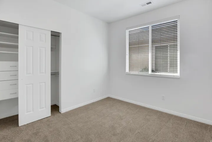 Neutral-toned carpet flooring, a window with horizontal blinds, built-in closet shelving, and white sliding closet doors with paneled detailing