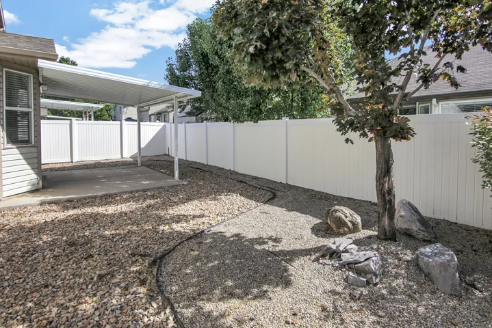 Carport structure with a concrete pad, gravel landscaping, white privacy fencing, and mature trees
