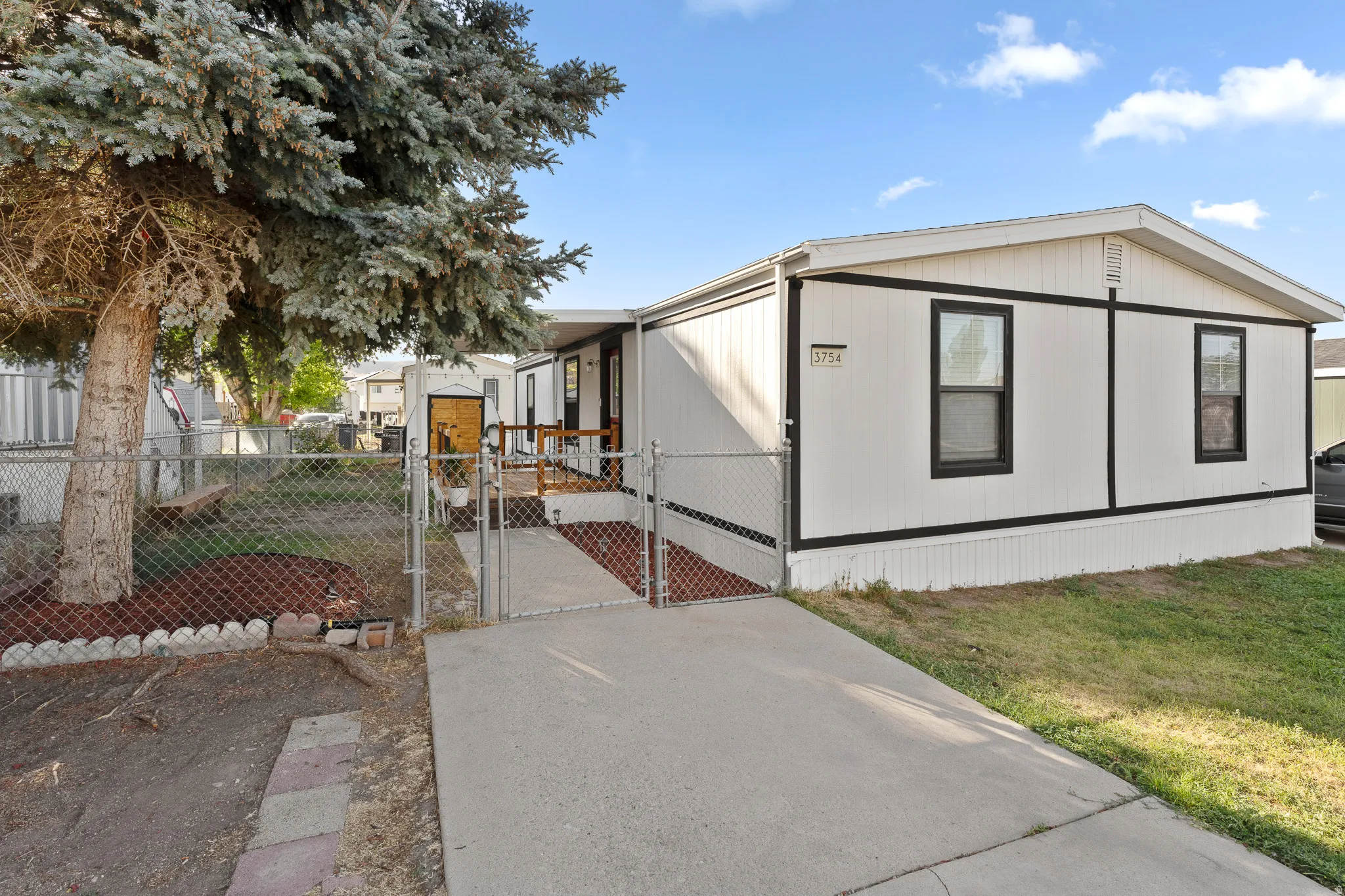 White exterior with black trim, concrete driveway, front entrance ramp, chain-link fence, and mature tree