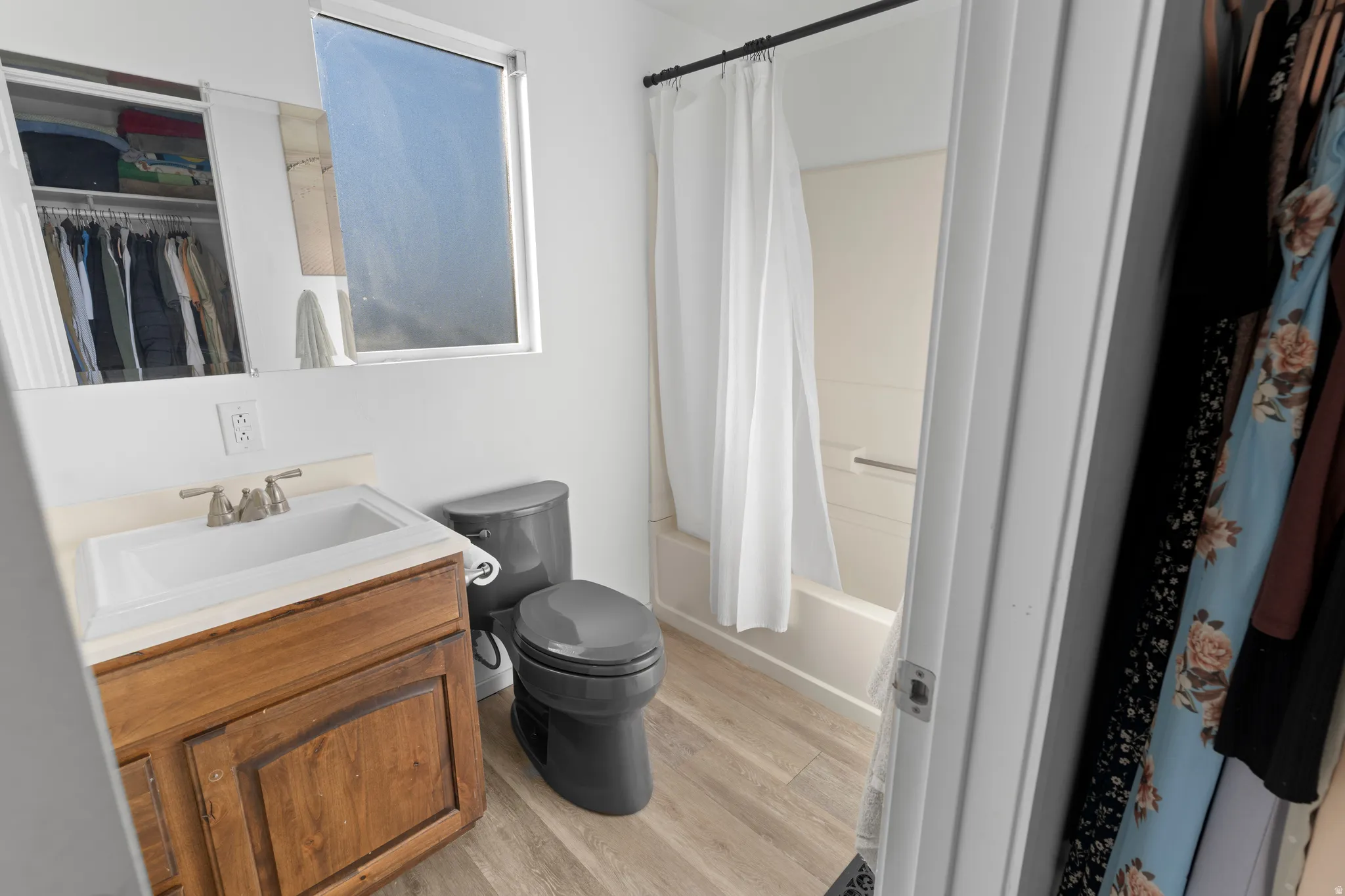 Wood-finish flooring, single-pane window, dark-toned toilet, vanity with integrated sink, and a shower-tub combination