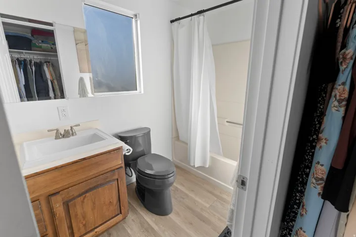 Wood-finish flooring, single-pane window, dark-toned toilet, vanity with integrated sink, and a shower-tub combination