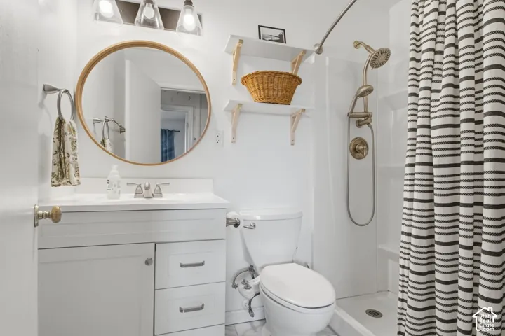 White vanity with brushed nickel hardware, circular mirror with wood-tone frame, triple light fixture, white shower surround, and a shower with a flexible hose attachment