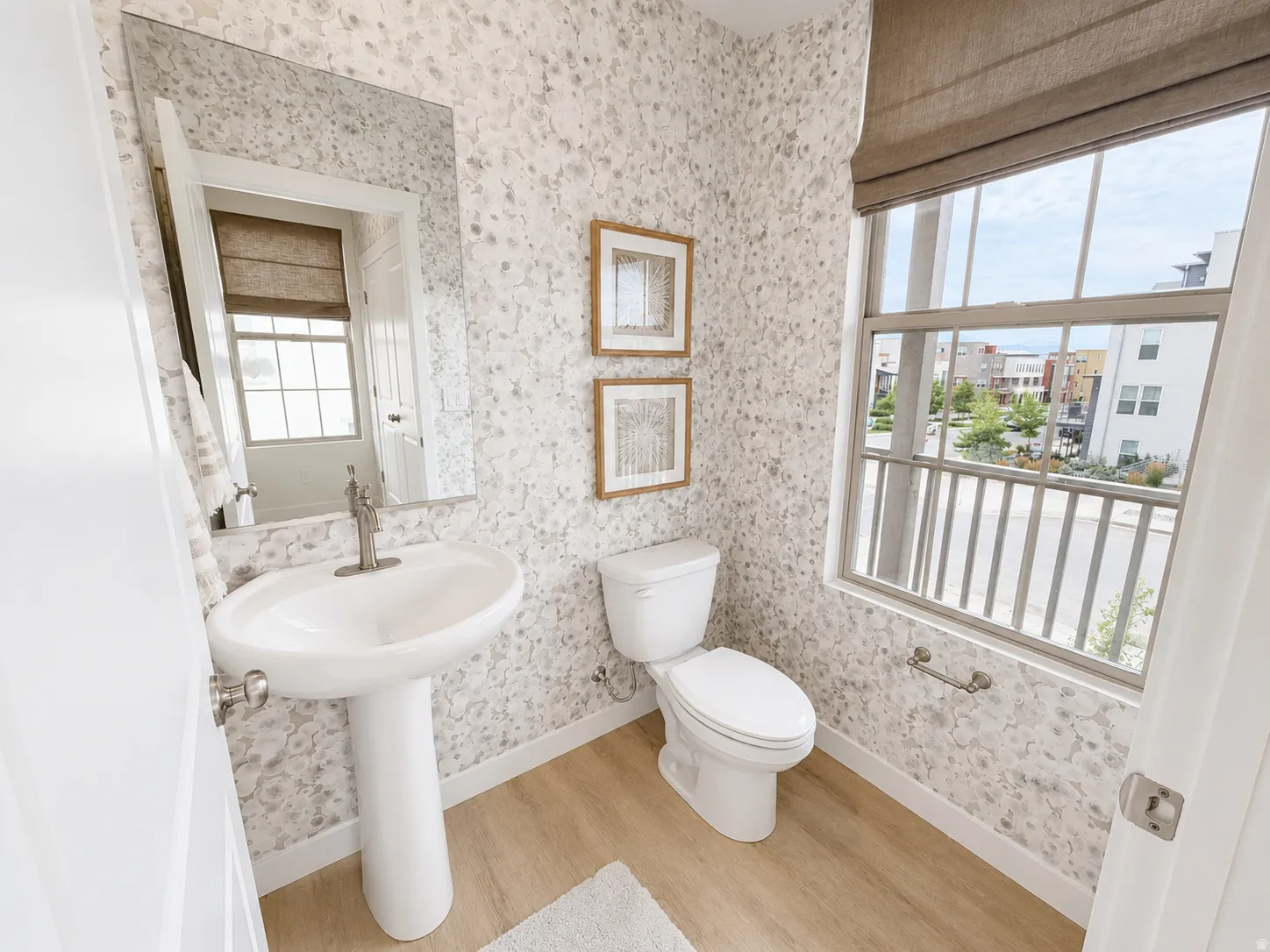 Pedestal sink with a brushed nickel faucet, wood-finish flooring, and a window with a roman shade