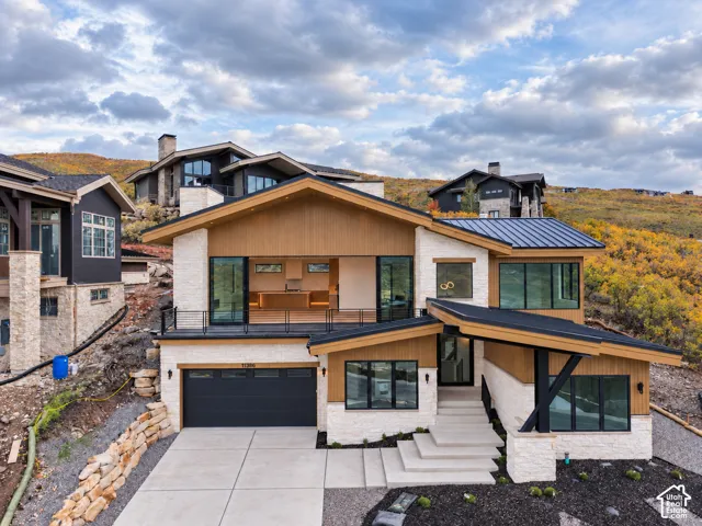 Modern architectural exterior featuring a combination of light-colored stone and wood-panel accents, a dark metal roof, large windows, and an integrated garage
