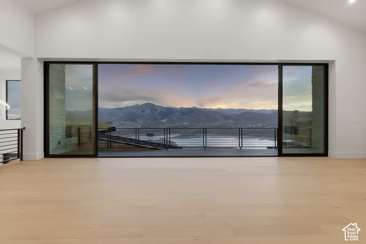 Vaulted ceilings, wood-finish flooring, and expansive multi-panel glass sliding doors opening to a deck featuring a metal railing with mountain and water scenery