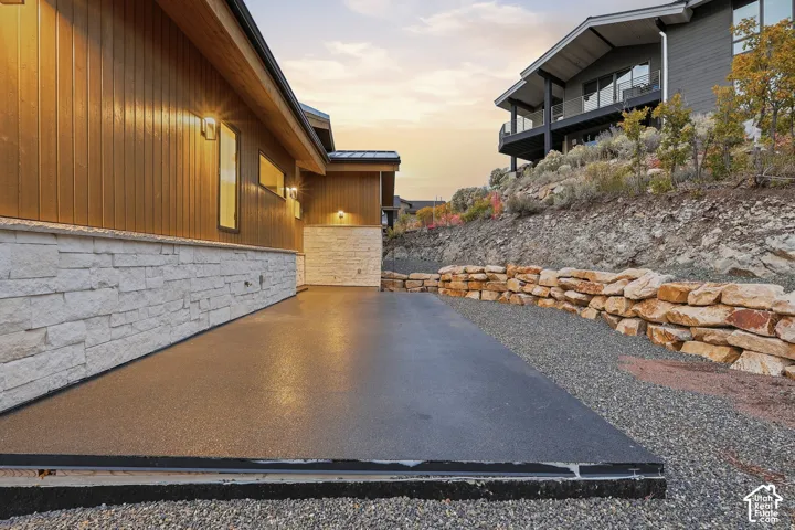 Exterior featuring vertical wood siding, white stone masonry, a concrete patio, and a tiered rock retaining wall