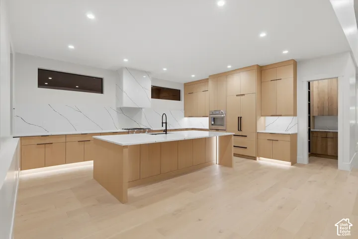 Kitchen featuring light wood-finish cabinetry, waterfall-edge island, patterned stone backsplash, built-in stainless steel appliances, and wood-finish flooring
