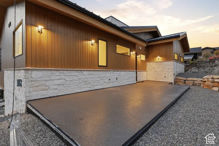 Contemporary exterior featuring vertical siding, stacked-stone masonry, expansive concrete patio, and wall-mounted exterior lighting
