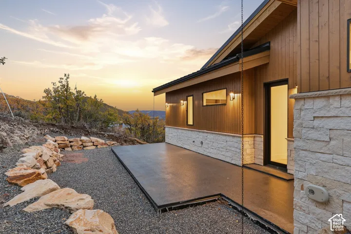 Exterior featuring vertical wood siding, light-colored stacked stone masonry, concrete patio, and a natural rock retaining wall