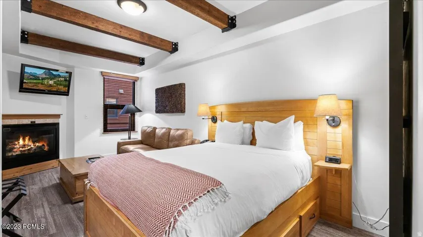 Exposed ceiling beams, wood-finish flooring, built-in fireplace, wall-mounted television, and a window with blinds