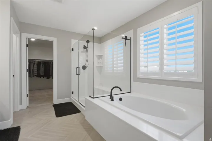Soaking tub with matte black fixtures, glass-enclosed shower, plantation shutters, and light-toned tile flooring