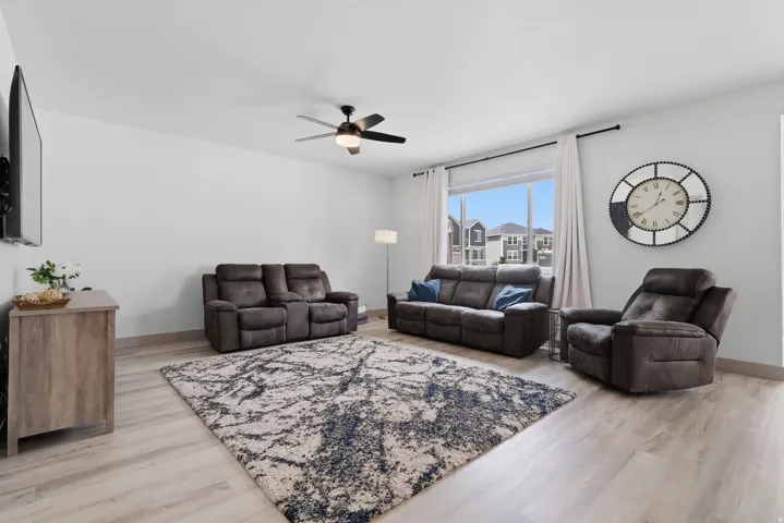 Spacious living area featuring light wood-finish flooring, a contemporary ceiling fan, and a large window with white drapery
