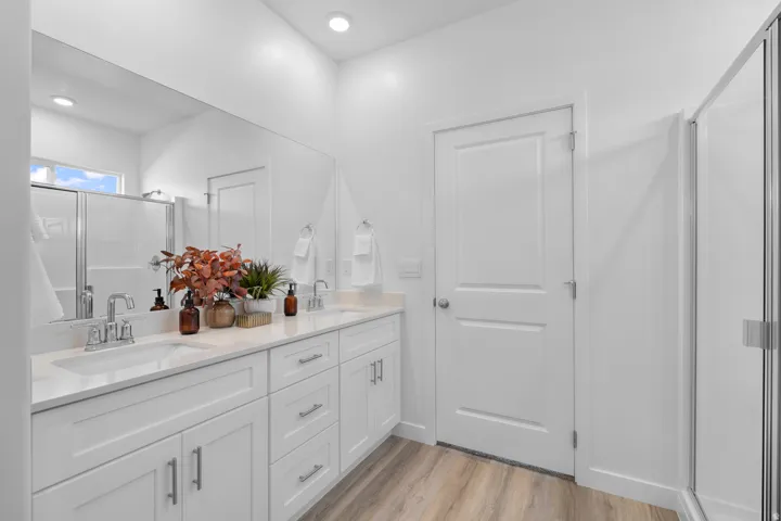 Dual vanity with undermount sinks and chrome fixtures, extensive wall mirror, white shaker-style cabinetry, wood-finish flooring, and a glass-enclosed shower