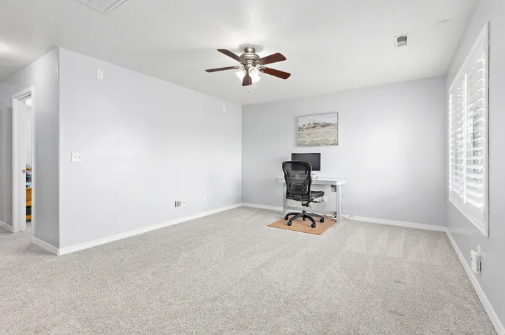 Plush carpeting, ceiling fan with light fixture, window with white plantation shutters, and light gray wall paint