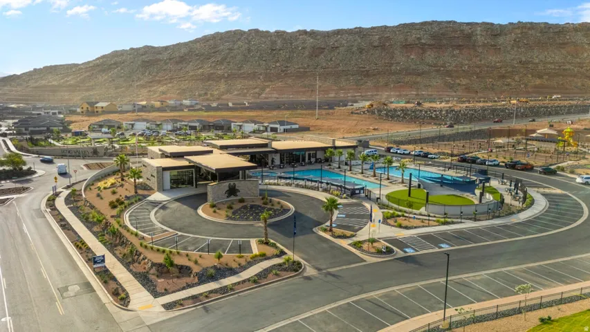 Community entrance featuring a multi-lane roundabout, mature palm trees, and a modern clubhouse with an outdoor swimming pool, integrated splash pad, and surrounding hardscaping