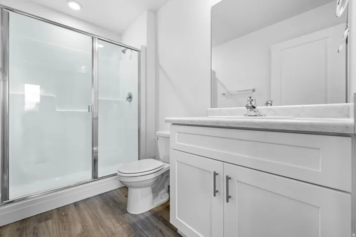 Walk-in shower with sliding glass doors, white vanity with integrated sink, chrome faucet, wall-mounted mirror, and wood-finish flooring