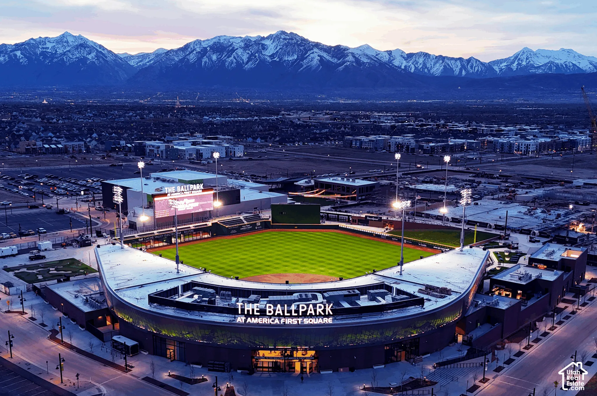 Ballpark stadium with an illuminated field and surrounding structures, set against a backdrop of snow-capped mountains and a developed area