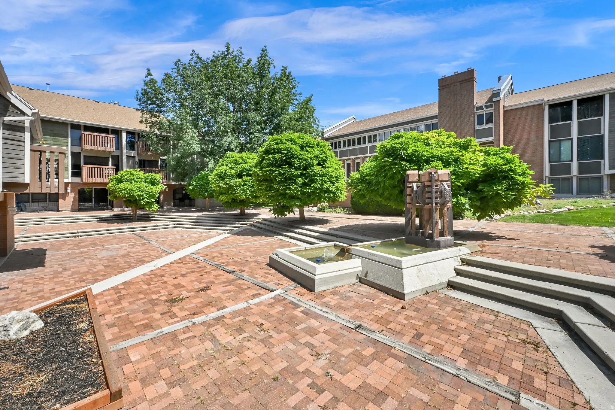 Expansive brick courtyard featuring mature trees, concrete steps, and a multi-tiered water feature with a central sculpture