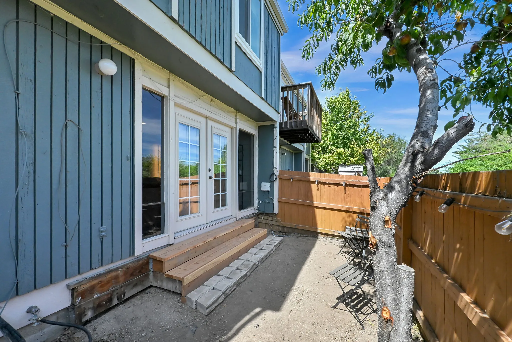 Private outdoor patio area with a wood fence & bistro lights, mature shade tree, and exterior wood siding.