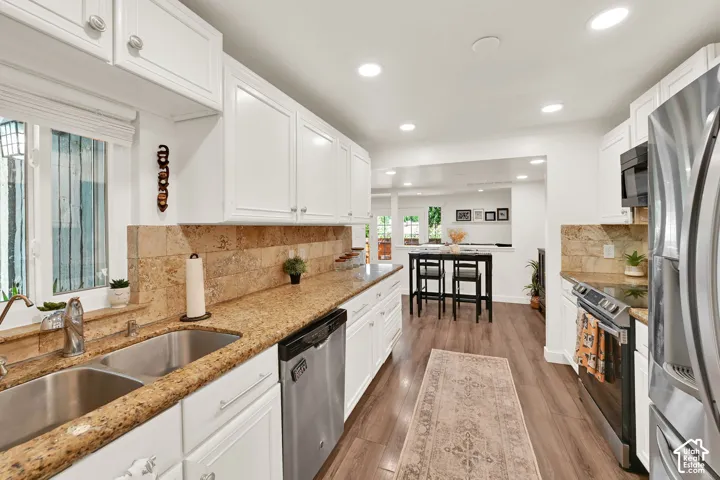 White cabinetry with brushed nickel hardware, light-toned stone countertops, a double basin stainless steel sink, tile backsplash, and recessed ceiling lighting