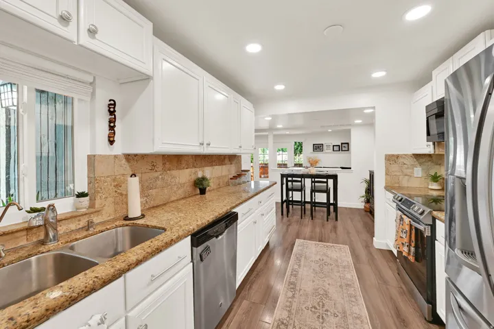 White cabinetry with brushed nickel hardware, light-toned stone countertops, a double basin stainless steel sink, tile backsplash, and recessed ceiling lighting