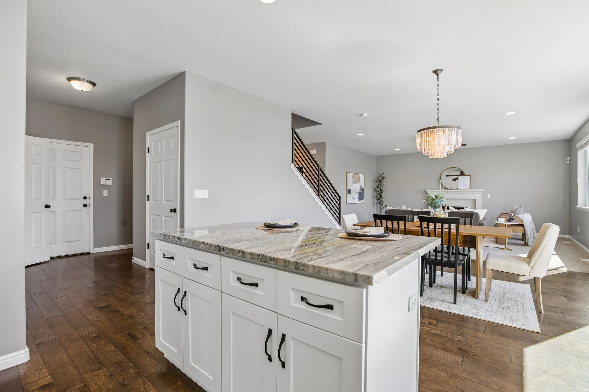 Kitchen island with stone countertop and white cabinetry, wood-finish flooring, staircase with black metal railing, dining area with chandelier, and living room with fireplace