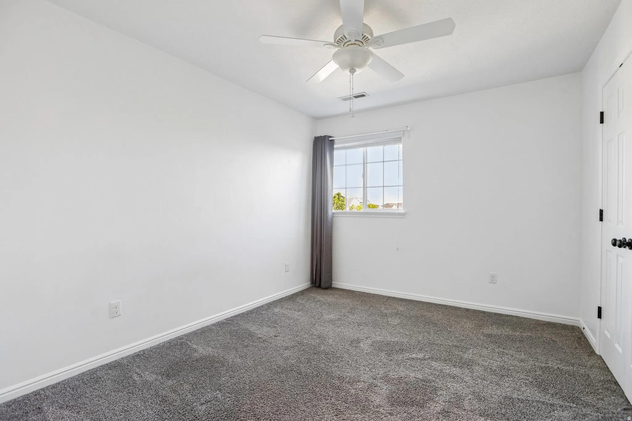 Neutral wall paint, ceiling fan with integrated lighting, window with white trim, gray textured carpeting, and a white paneled door with black hardware