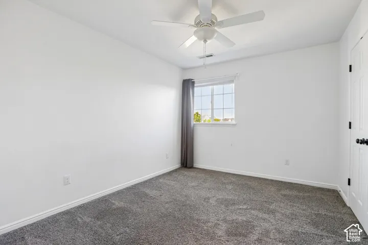 Neutral wall paint, ceiling fan with integrated lighting, window with white trim, gray textured carpeting, and a white paneled door with black hardware