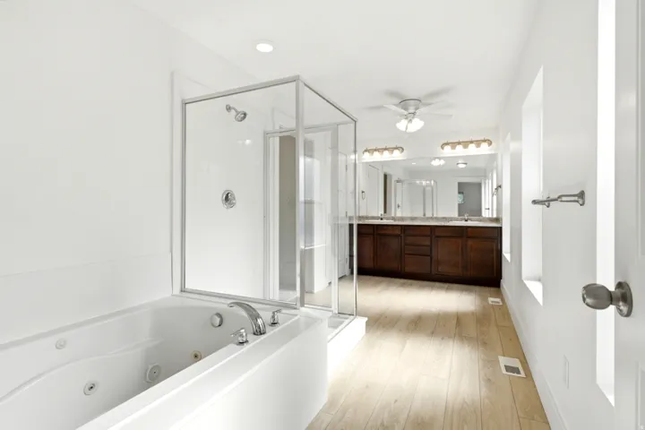 Soaking tub, glass-enclosed shower, dual vanity with dark wood cabinetry, integrated lighting, and wood-finish flooring