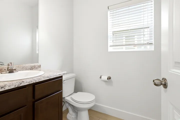 Window with horizontal blinds, white toilet, wood-finish vanity with an integrated sink, and a speckled countertop