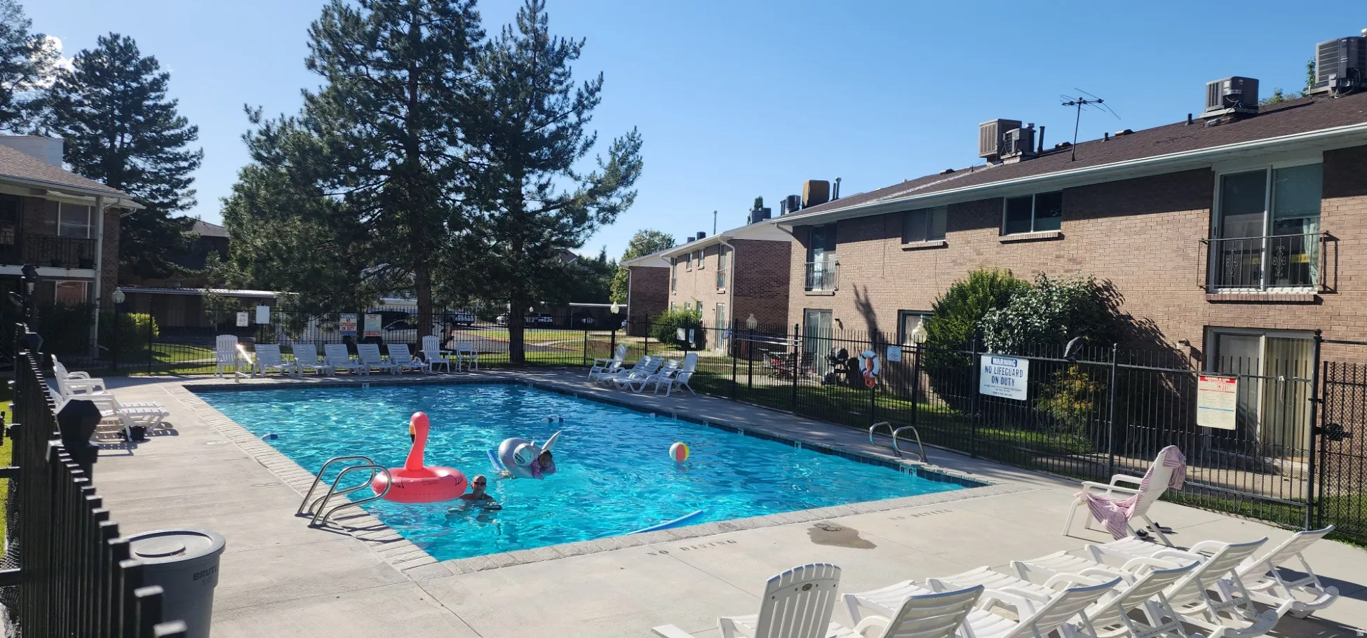 Outdoor swimming pool with a surrounding paved deck, brick exterior buildings, and mature trees