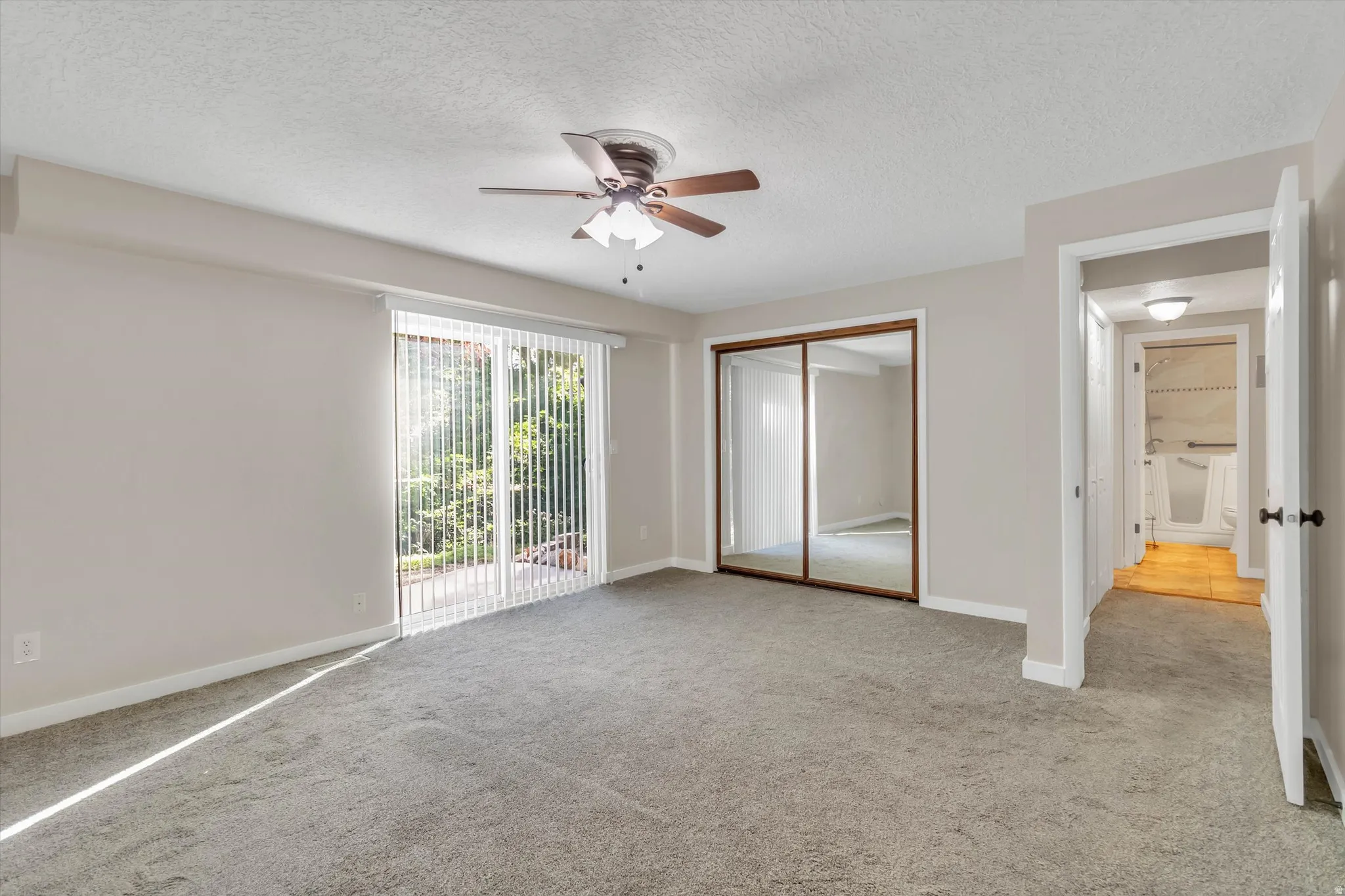 Spacious room with plush carpeting, sliding glass door with vertical blinds, mirrored closet doors, and a ceiling fan
