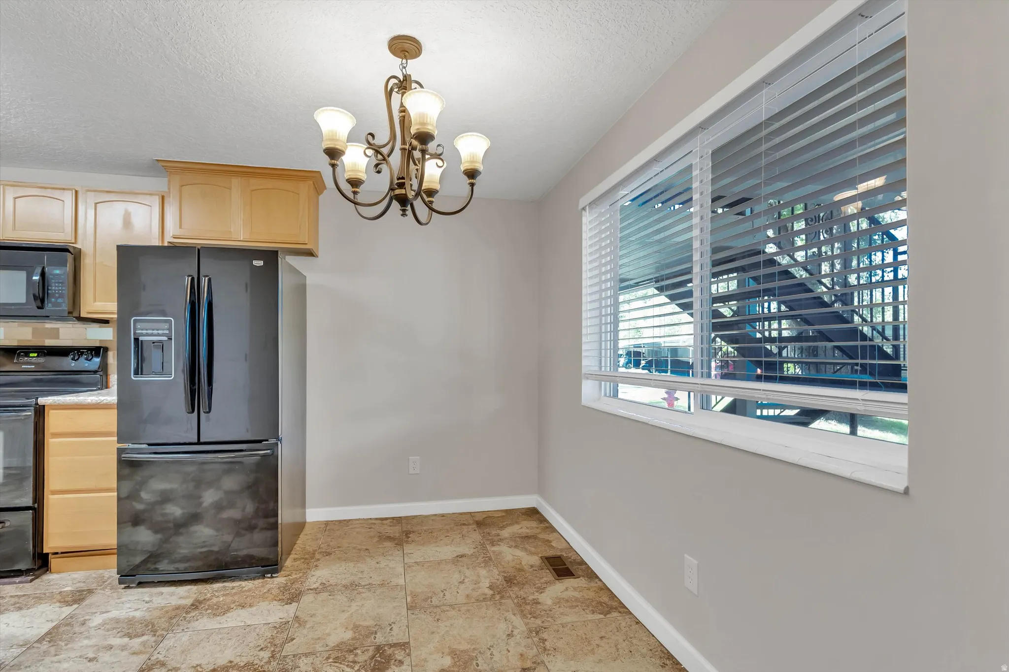Tile flooring, light wood-finish cabinetry, black French door refrigerator, built-in microwave, and a five-light chandelier