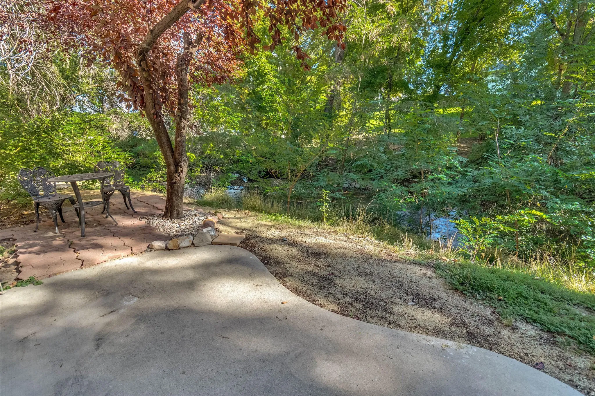 Concrete patio with an adjoining paver section under a mature tree, featuring a natural creek beyond