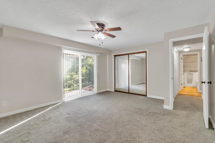 Spacious room with plush carpeting, sliding glass door with vertical blinds, mirrored closet doors, and a ceiling fan