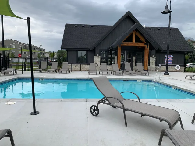Community swimming pool with a concrete deck, multiple lounge chairs, and a contemporary clubhouse featuring stone accents and dark siding