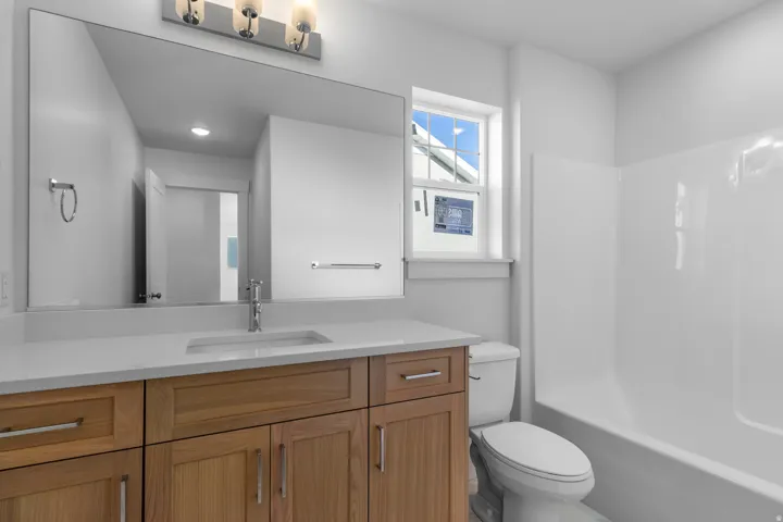 White vanity countertop, wood-finish cabinetry with silver-tone hardware, rectangular undermount sink, contemporary wall-mounted light fixture, and white tub/shower combination