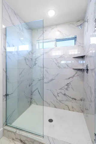 Shower enclosure featuring clear glass panels, marble-look wall tiles, a mosaic tile shower floor, polished chrome fixtures, and built-in shelves