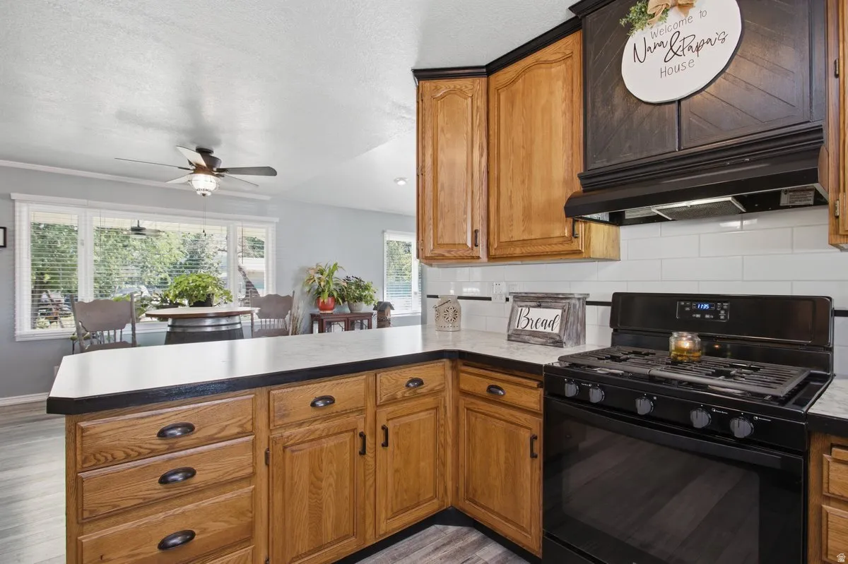 Wood-finish cabinetry, white Formica countertops, black gas range, subway tile backsplash, and light wood-finish laminate flooring