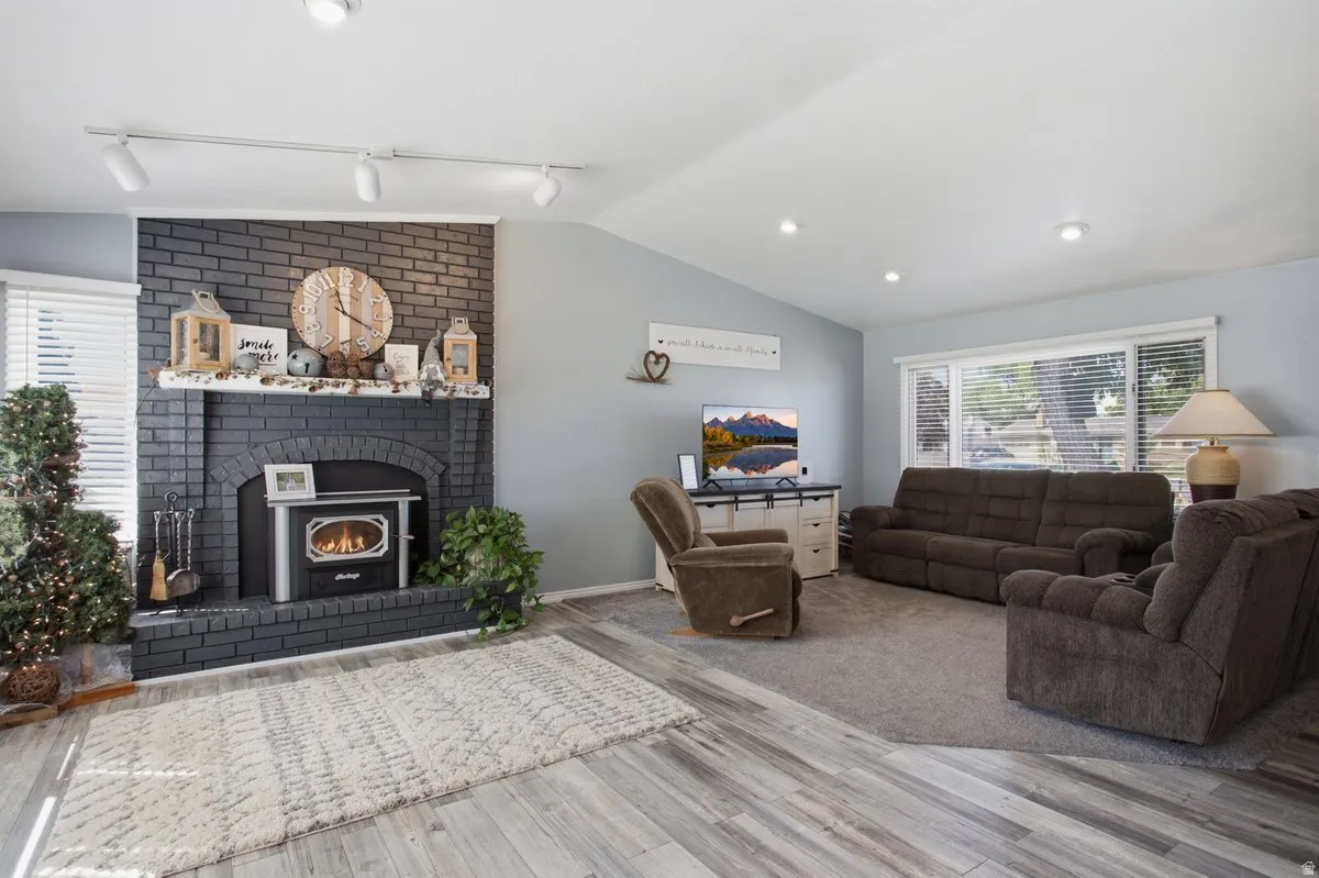 Vaulted ceiling with recessed lighting, black brick fireplace with a wood-burning stove, large windows with horizontal blinds, and wood-finish flooring