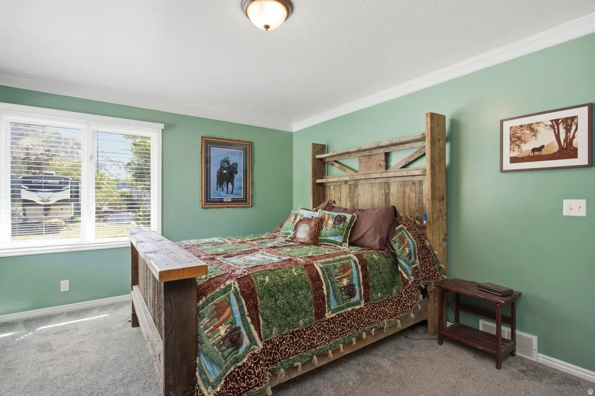 Double-paned window with horizontal blinds, wood-finish bed frame, carpet flooring, crown molding, and wall-mounted light fixture